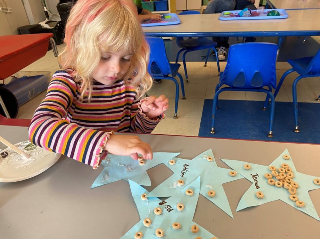 Photo of a young girl sitting at a table, gluing cheerios to a paper star.