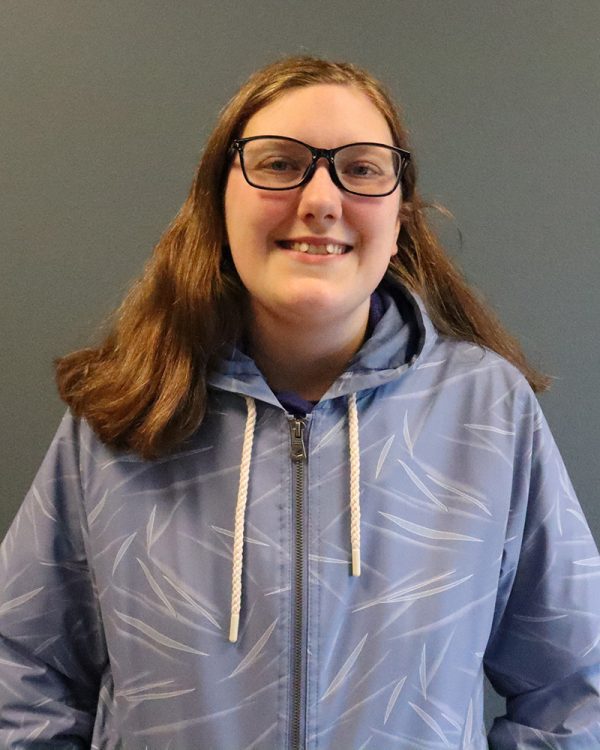 Photo of a young woman in a hoodie and glasses standing in front of a blank and smiling.